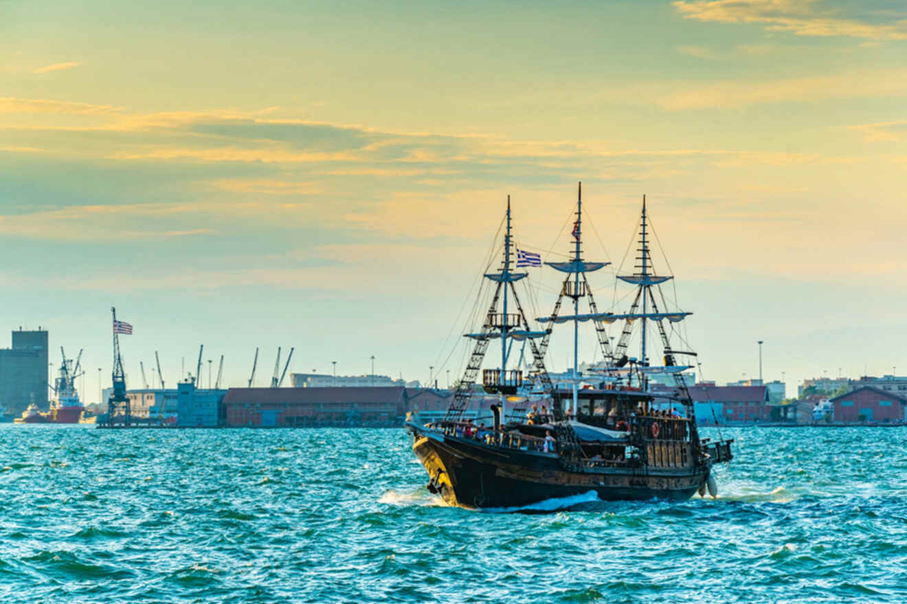 A vintage-style ship with three masts sails on a blue sea, with an industrial port and cranes visible in the background under a partly cloudy sky.