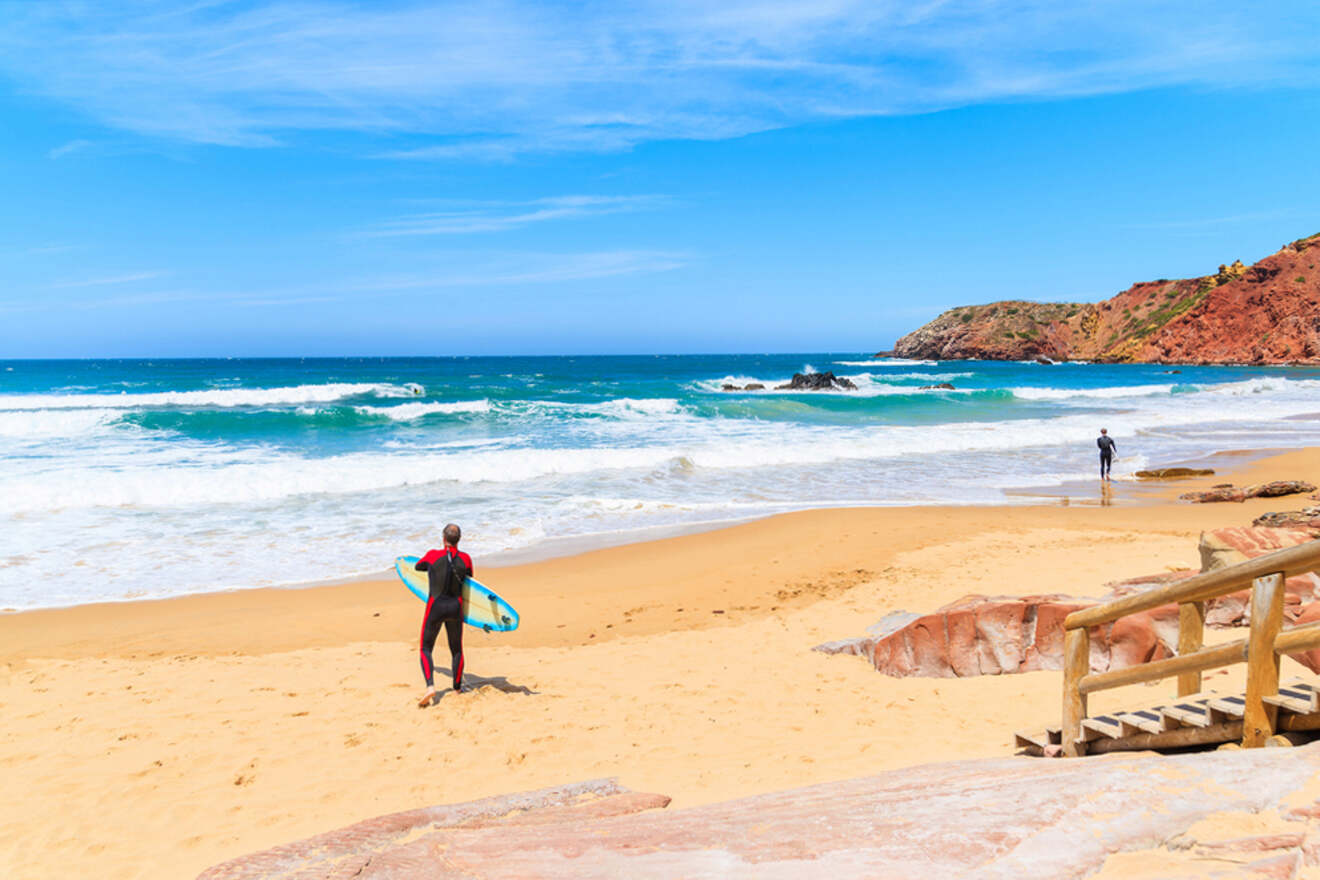 A surfer in a wetsuit holding a surfboard faces the ocean on a sandy beach in Lagos, Portugal, with another person nearby and rocky cliffs in the distance under a blue sky.