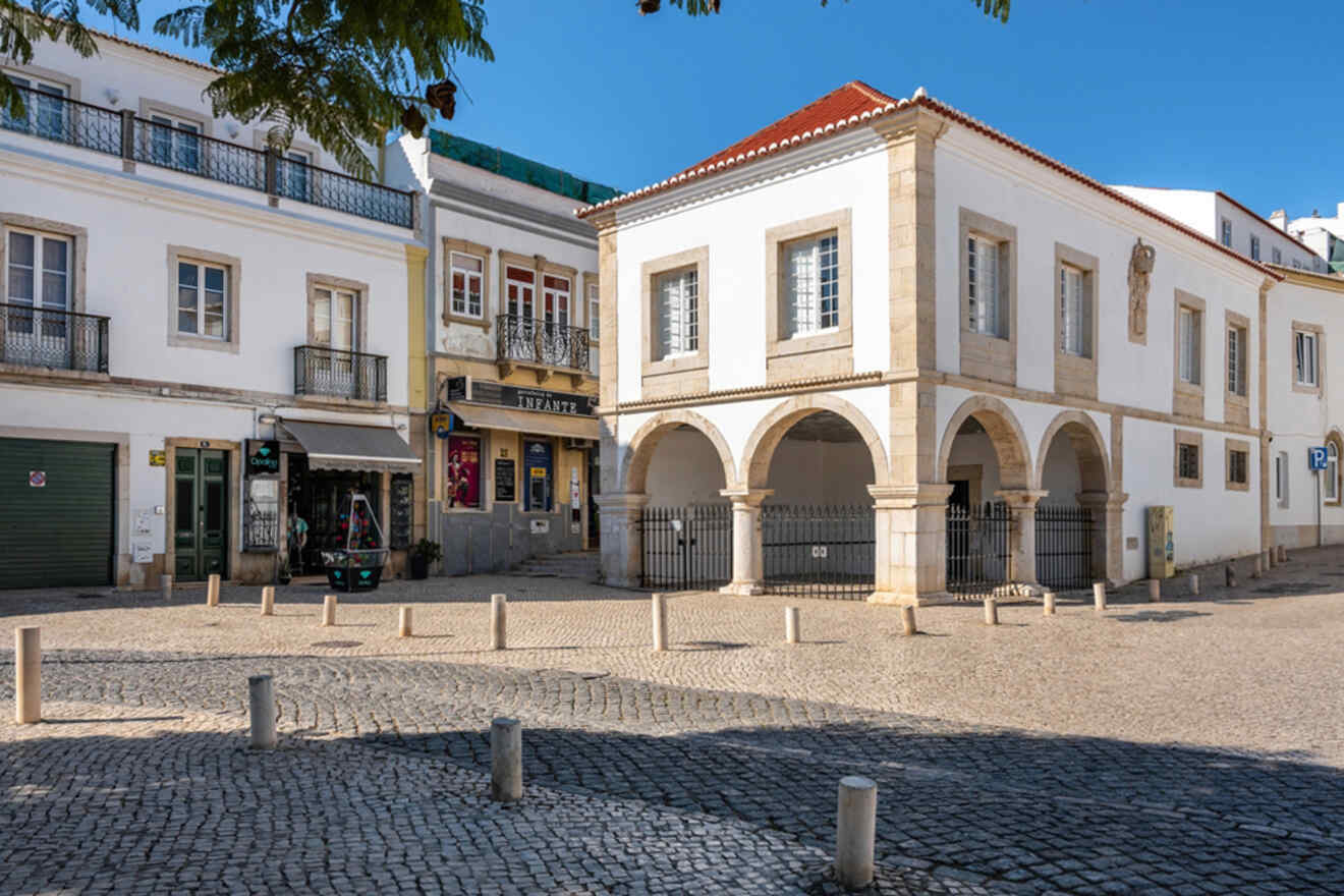 A cobblestone square in Lagos, Portugal, boasts a historic white building with elegant arches and columns. Nearby, a few shops with closed shutters and balconies sit under the clear blue sky.