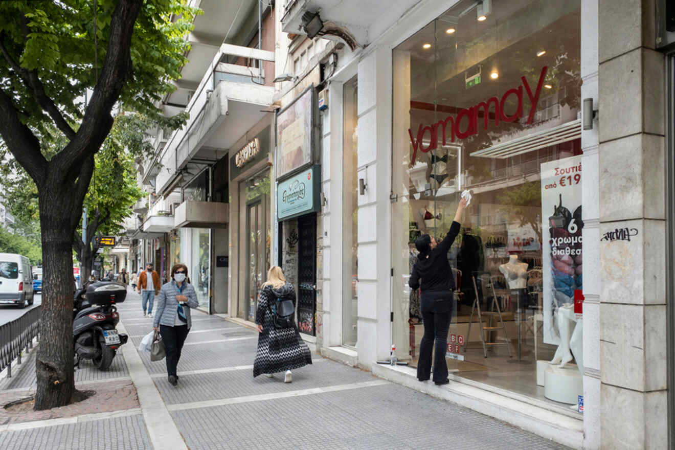 People walk past a storefront with mannequins and sale signs. A person adjusts a display inside. A scooter is parked nearby. Trees line the street.