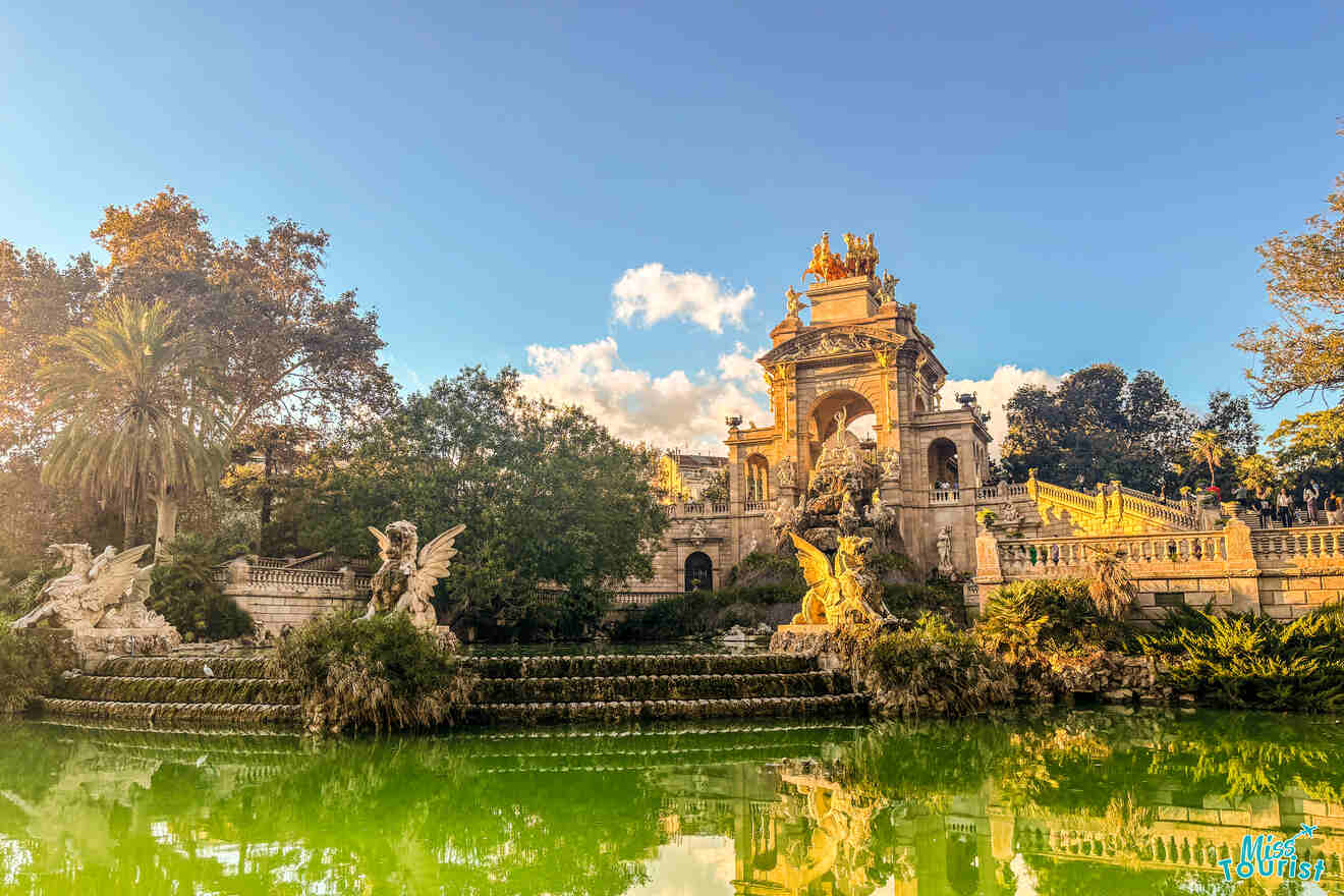 Fountain with stone statues and ornate archway surrounded by lush greenery under a clear blue sky.