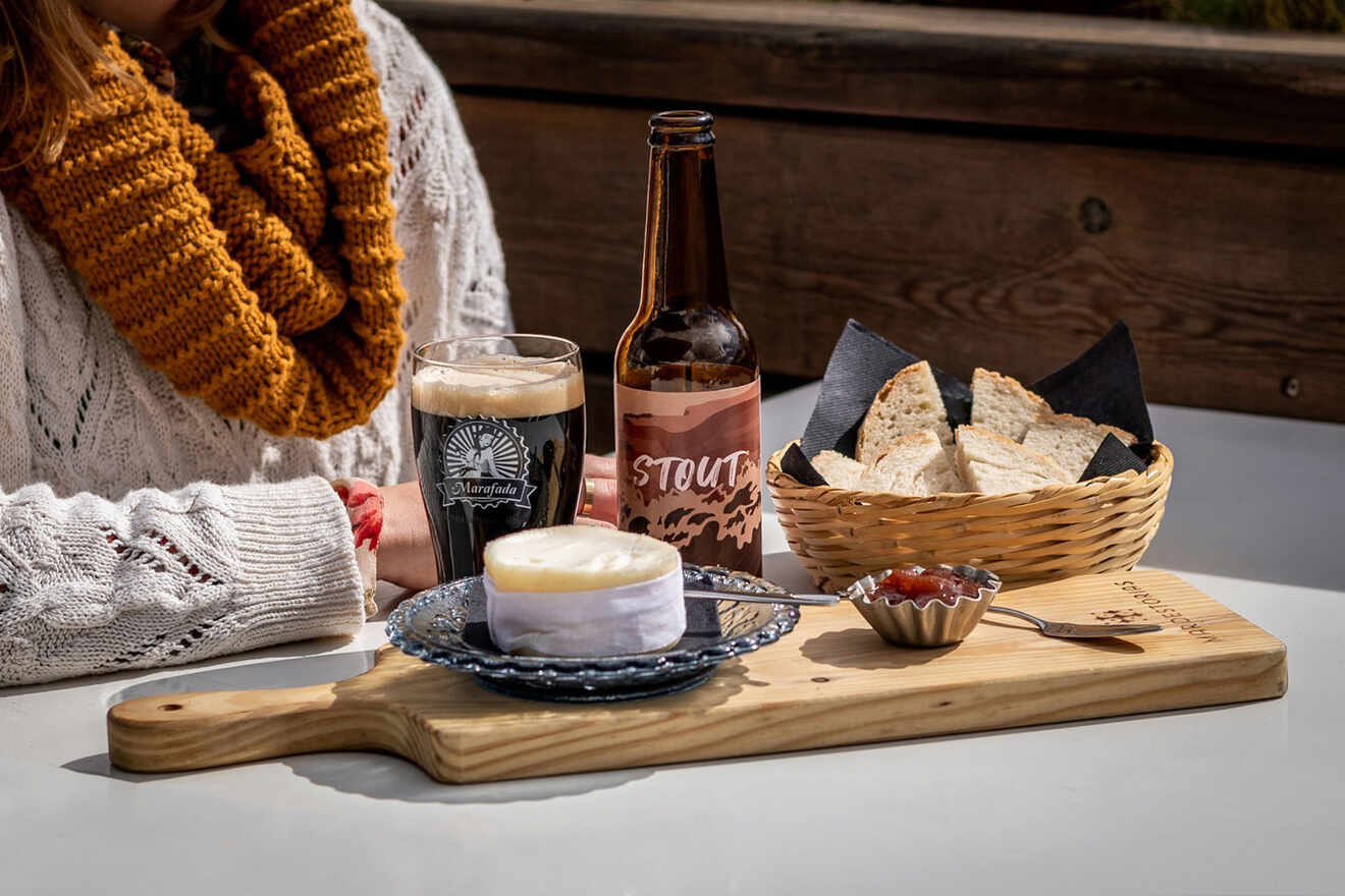 A woman sitting at a table with a wooden board holding cheese, crackers, and jam. A glass of dark beer and an unopened beer bottle are next to a basket of bread.