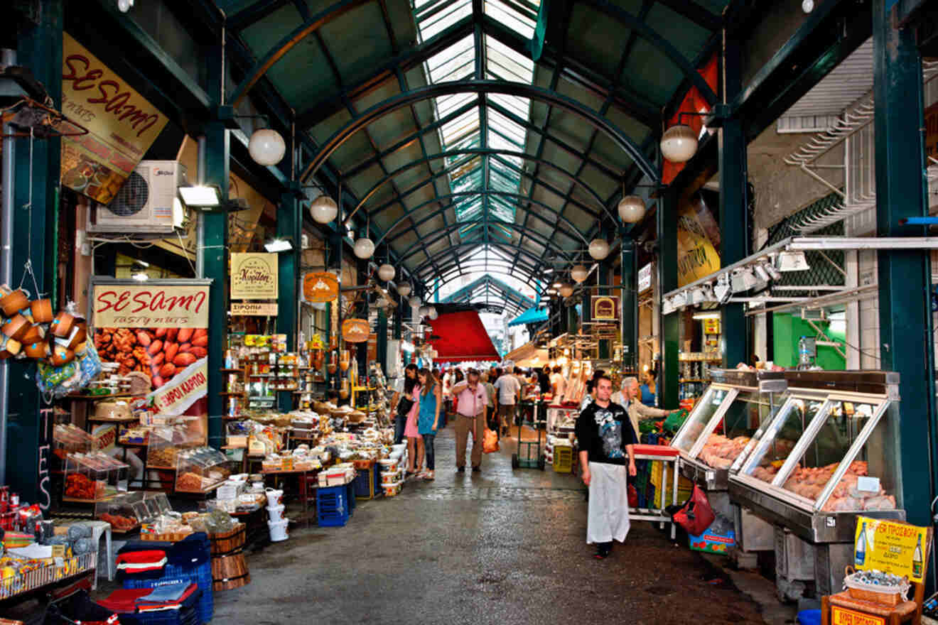 A bustling indoor market with various stalls selling food and goods. Shoppers and vendors walk along the covered aisle.