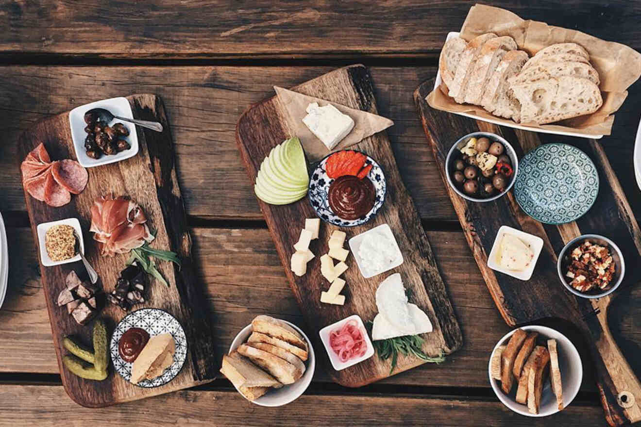 Wooden boards display a variety of cheeses, breads, olives, meats, dips, and sliced fruits on a rustic wooden table.