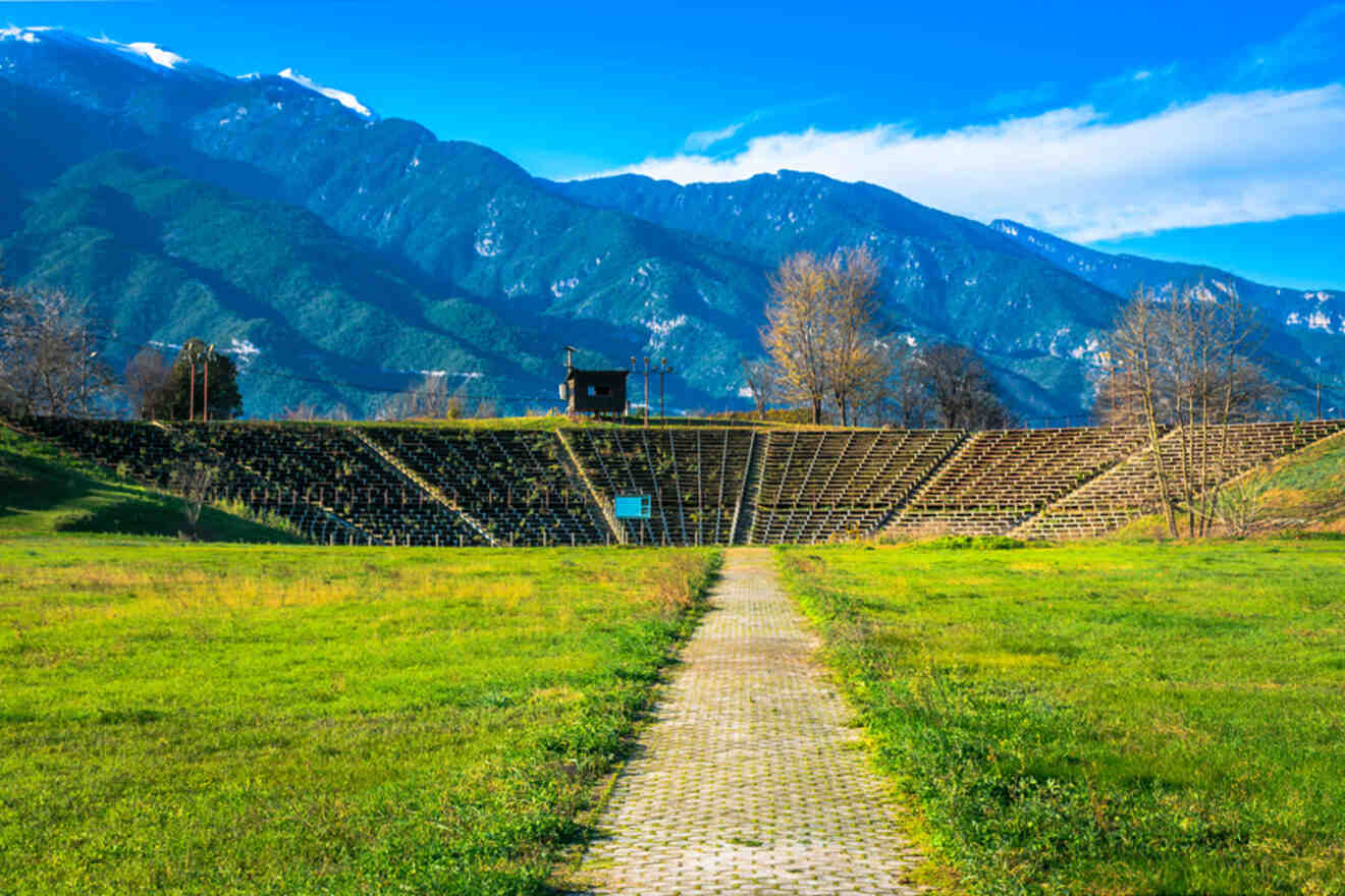 Ancient amphitheater with stone seating, surrounded by green grass and with mountains in the background under a clear blue sky.