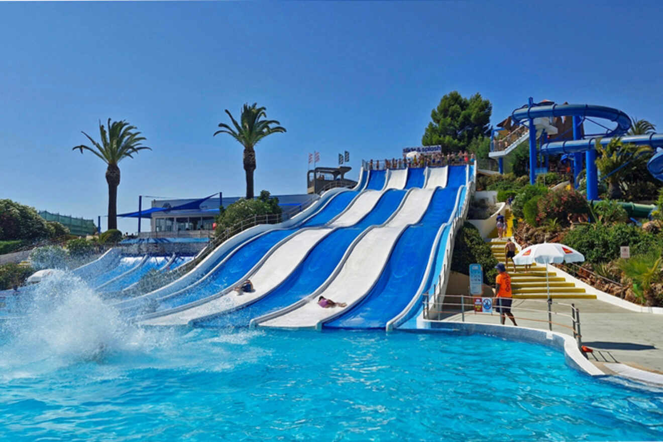 People in Lagos, Portugal, are sliding down multiple blue and white water slides into a large pool, with palm trees swaying against the clear sky in the background.