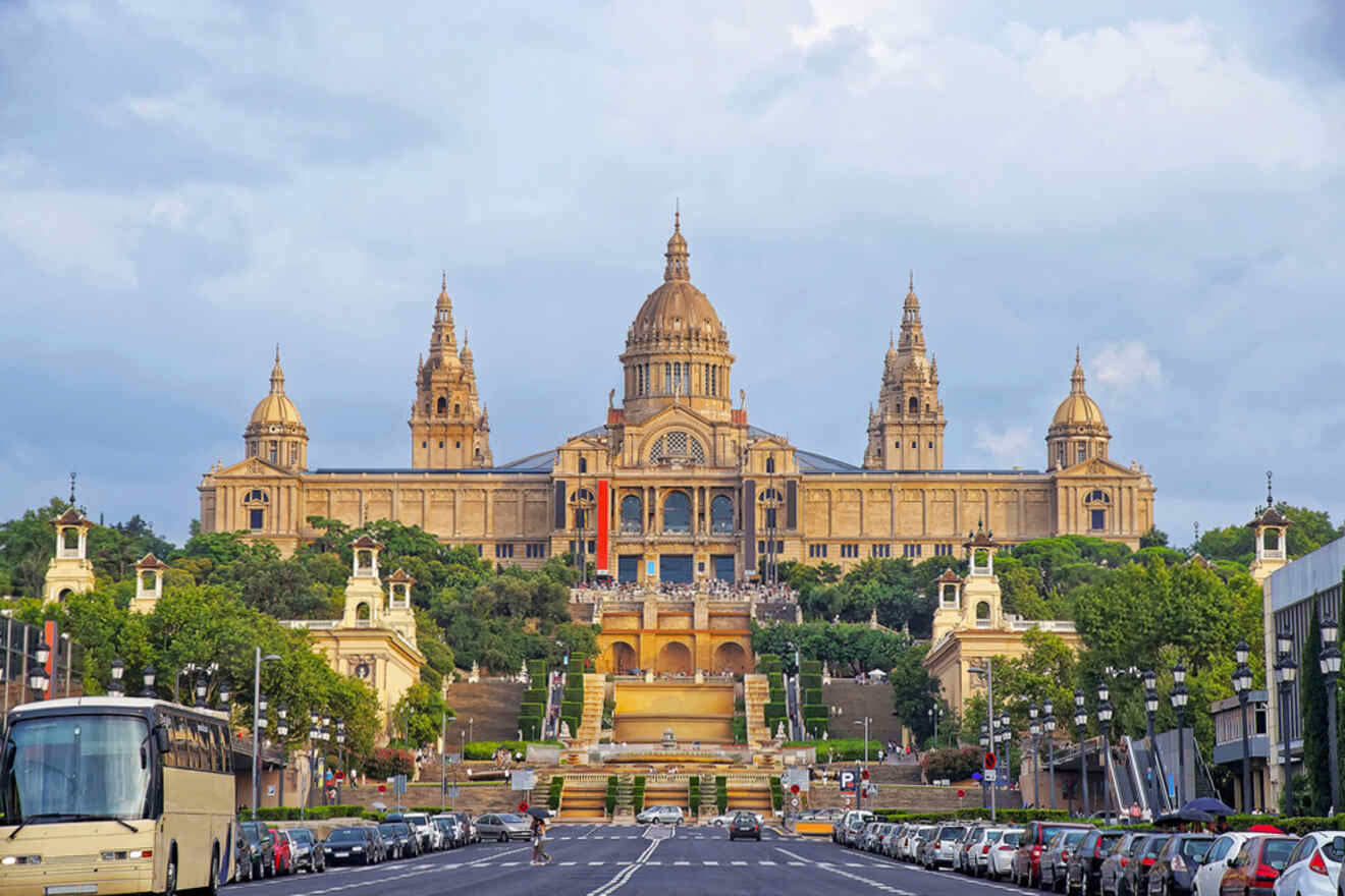 Grand neoclassical building with towers and domes, surrounded by greenery; parked cars and a road in the foreground.