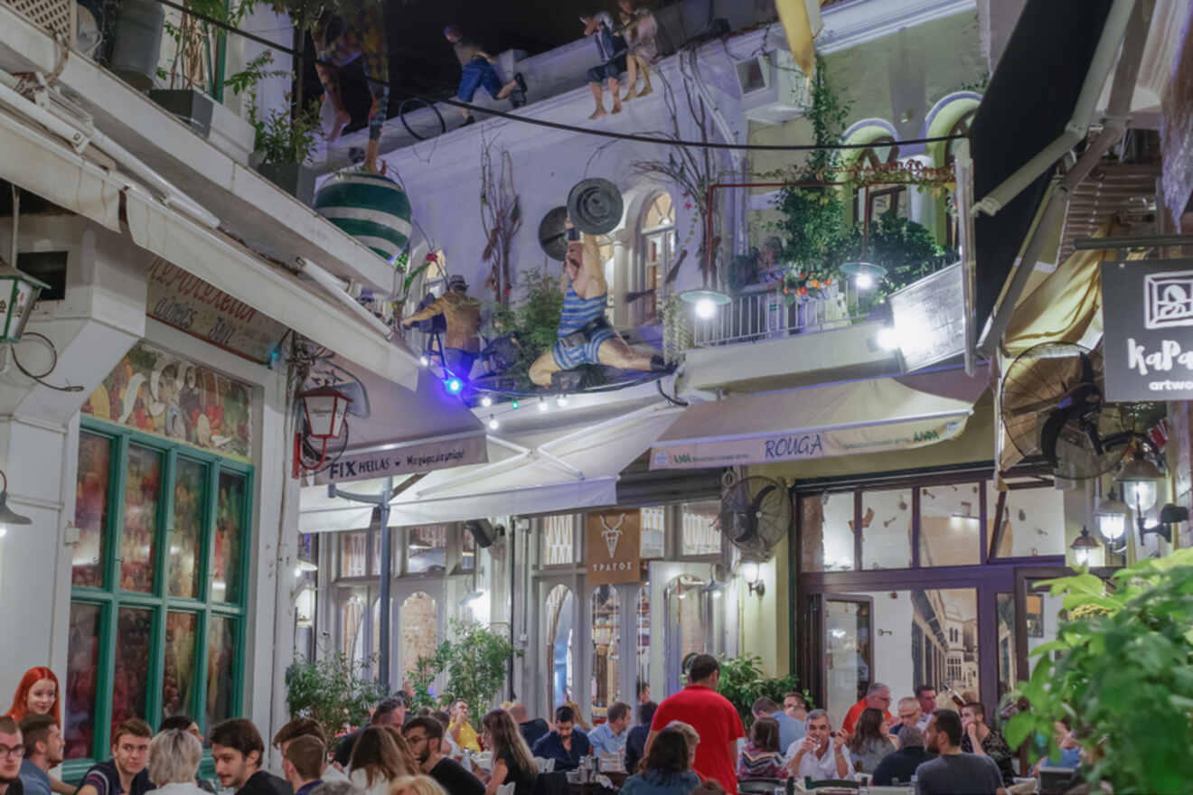 A busy outdoor restaurant with people dining under a balcony decorated with playful sculptures, including a man lifting weights.