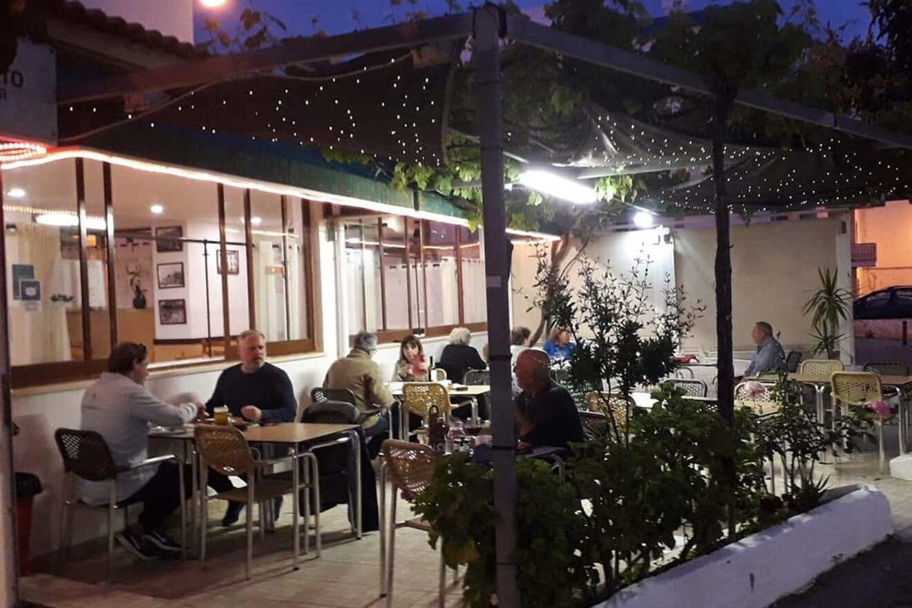 Outdoor café with several people seated at tables under a canopy, surrounded by plants, in evening light.