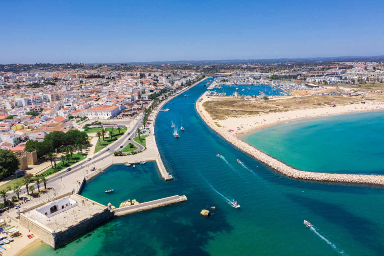 Aerial view of Lagos, Portugal, a coastal city where the river gracefully meets the sea, lined with boats and sandy beaches. The vibrant mix of urban and green areas basks under a clear blue sky.