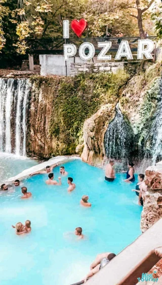 People enjoying a thermal pool near a waterfall, with a sign reading "I ♥ POZAR" in the background.