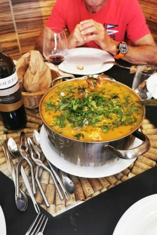 A pot of seafood stew garnished with herbs on a table, accompanied by a bottle of wine, empty glasses, bread, and cutlery. A person in a red shirt sits nearby with folded hands.