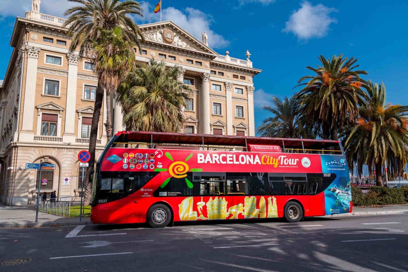 A red double-decker Barcelona City Tour bus parked by a historical building with palm trees and a clear blue sky.