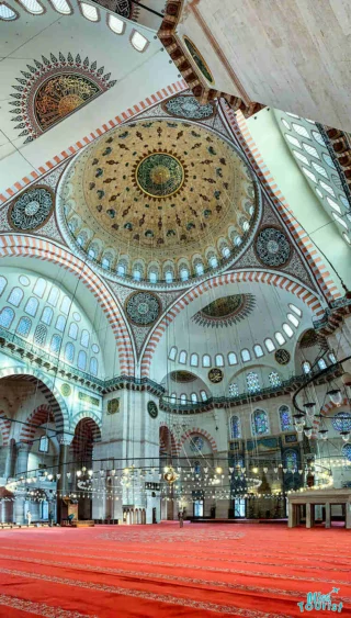 Interior of a grand mosque with ornate domed ceilings, intricate patterns, and red carpeted floors. Large chandeliers hang from above.