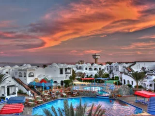 Resort with white buildings, a swimming pool, and palm trees under a colorful sunset sky.