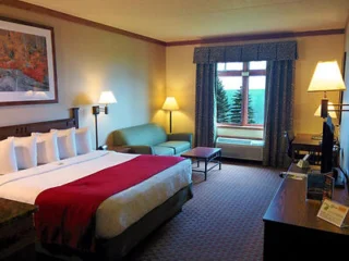 Hotel room with a king-sized bed, red throw blanket, sofa, desk, TV, and window with a view. Decor includes lamps and a framed picture above the bed.