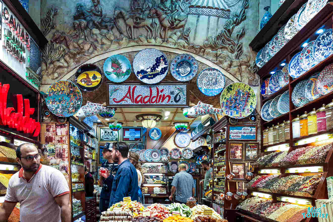 A bustling spice and souvenir shop with colorful pottery, plates, and various spices on display. Several people browse the items in the well-lit, ornate interior.