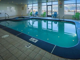 Indoor swimming pool with blue water, surrounded by a tiled floor. There are colorful chairs and large windows with a view of a hilly landscape outside.