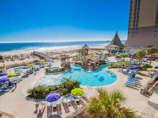 Aerial view of a beach resort with a large pool, sun loungers, umbrellas, and a wooden boardwalk leading to the ocean. A tall building stands to the right. Bright, sunny day.