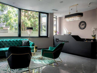 Modern reception area with a dark counter, wall clock, and two chandeliers. Features green sofa and chairs around a glass coffee table, large windows, and potted plants.