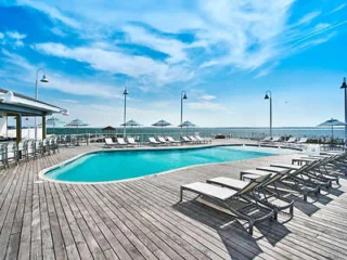 Outdoor pool area with wooden deck, lounge chairs, and umbrellas overlooking the ocean under a clear blue sky.