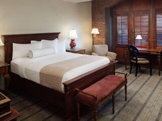 Hotel room with a large bed, wooden headboard, bedside tables with lamps, an upholstered bench, armchair, desk, and wooden shutters on a brick wall.