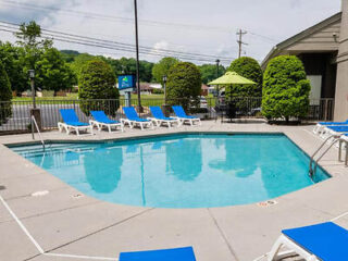 Outdoor swimming pool with blue lounge chairs arranged along the sides. Green umbrellas and landscaped bushes surround the area.