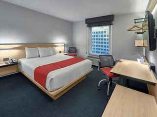 Hotel room with a king-size bed, red accent blanket, desk with chair, mounted TV, and a window with a city view.