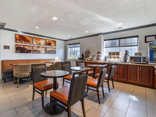 A small cafe-style seating area with round tables, chairs, a coffee station, and artwork on the walls. Large windows provide natural light.