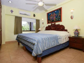 Bedroom with a large wooden bed, colorful wall art, two lamps on bedside tables, a ceiling fan, and a window with curtains.