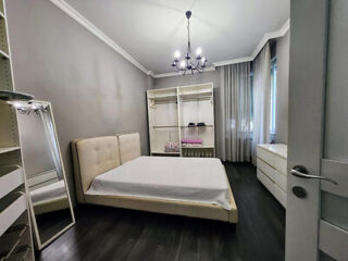 Minimalist bedroom with a beige bed, open wardrobe, standing mirror, and chandelier. Gray walls and dark wood flooring complement the space.