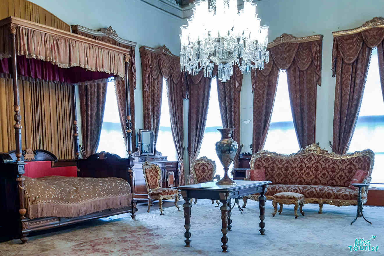 Ornate period room with canopy bed, elegant chandelier, patterned sofa, armchairs, and a decorated table with vase.