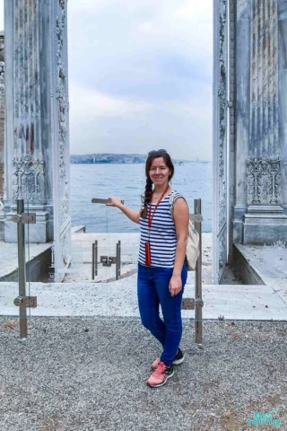 author of the post stands in front of an open marble gate by a waterfront, wearing a striped shirt, jeans, and pink shoes, holding the gate's post with one hand. The sky is overcast.