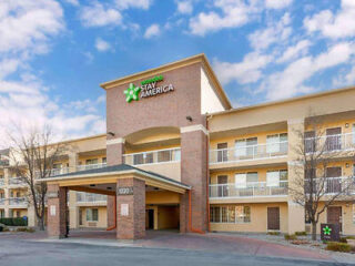 Three-story Stay America hotel entrance with a brick facade and covered drop-off area on a clear day.