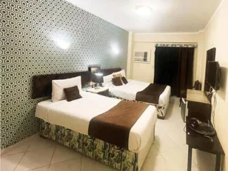 Hotel room with two single beds, decorative wall pattern, wall-mounted TV, desk, and curtains partially open.