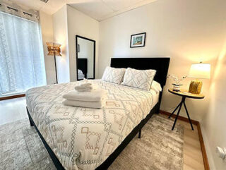 Bedroom with a neatly made bed, patterned bedding, a mirror on the wall, a table lamp on a round side table, and a small wall art piece. White and neutral tones dominate the decor.