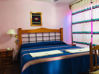 A neatly made bed with a blue and pink striped cover in a room with pink walls, a wooden headboard, wall-mounted lamp, framed artwork above, and a window with striped curtains.