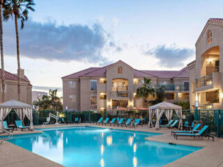 Pool area surrounded by lounge chairs and cabanas, with a three-story building and palm trees in the background under a partly cloudy sky.