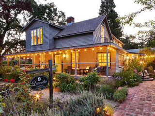 A charming two-story house with a wraparound porch, illuminated by string lights. Surrounded by lush gardens and a brick path, the scene exudes a cozy, welcoming atmosphere.