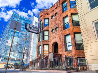 Historic red brick building with a turret next to a modern glass structure on a city street. A sign reads "Old Chicago Inn." Bright blue sky with clouds in the background.