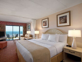 Hotel room with a king-sized bed, two lamps on nightstands, framed art on walls, and a balcony with a sea view.