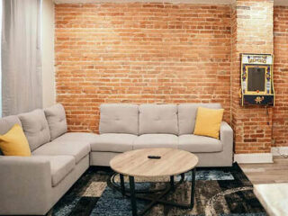 A cozy living room with a gray sectional sofa, a round wooden coffee table, and an arcade machine against a brick wall.