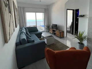 A living room with a blue sectional sofa, orange armchair, small TV on a stand, and a view of the ocean through sliding glass doors. A small rug and plants decorate the space.