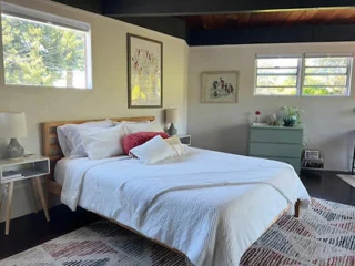 A bedroom with a neatly made bed, white bedding, two bedside tables with lamps, a dresser with decor, and a large window providing natural light.