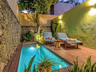 Small courtyard with a narrow swimming pool, two lounge chairs, potted plants, and wall lights.