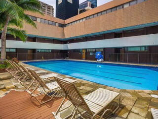 Outdoor swimming pool surrounded by lounge chairs, with a palm tree to the left and a multi-story building in the background.