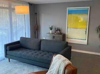 A modern living room with a gray sofa, a large abstract painting on the wall, a tall sculpture, and a console with bottles. A curtain and a floor lamp are visible.