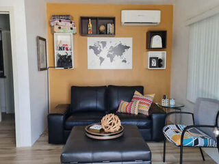 A living room with a black sofa, patterned cushions, wall shelves, a world map, and a round ottoman table. The wall is orange and decorated with various items.