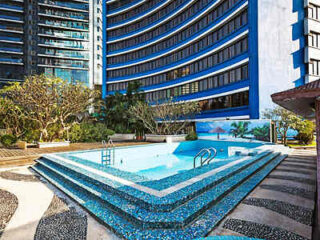 Empty outdoor swimming pool surrounded by modern high-rise buildings and greenery under a clear sky.