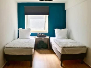 A small room with two single beds, a central window with a dark blind, and a small table between the beds. The walls are white and one is painted teal.